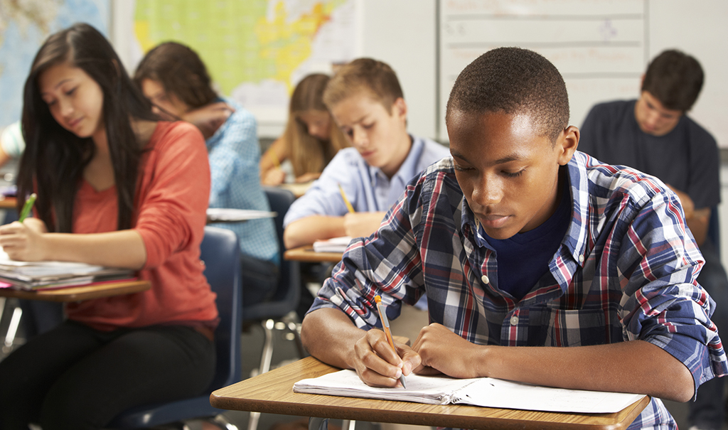 Teenage African American male wearing a plaid button-up shirt sitting at a desk in class. He is writing in a notebook on his desk using a pencil.  Around him are his classmates, also writing on their papers.  They are all concentrating on their own work.